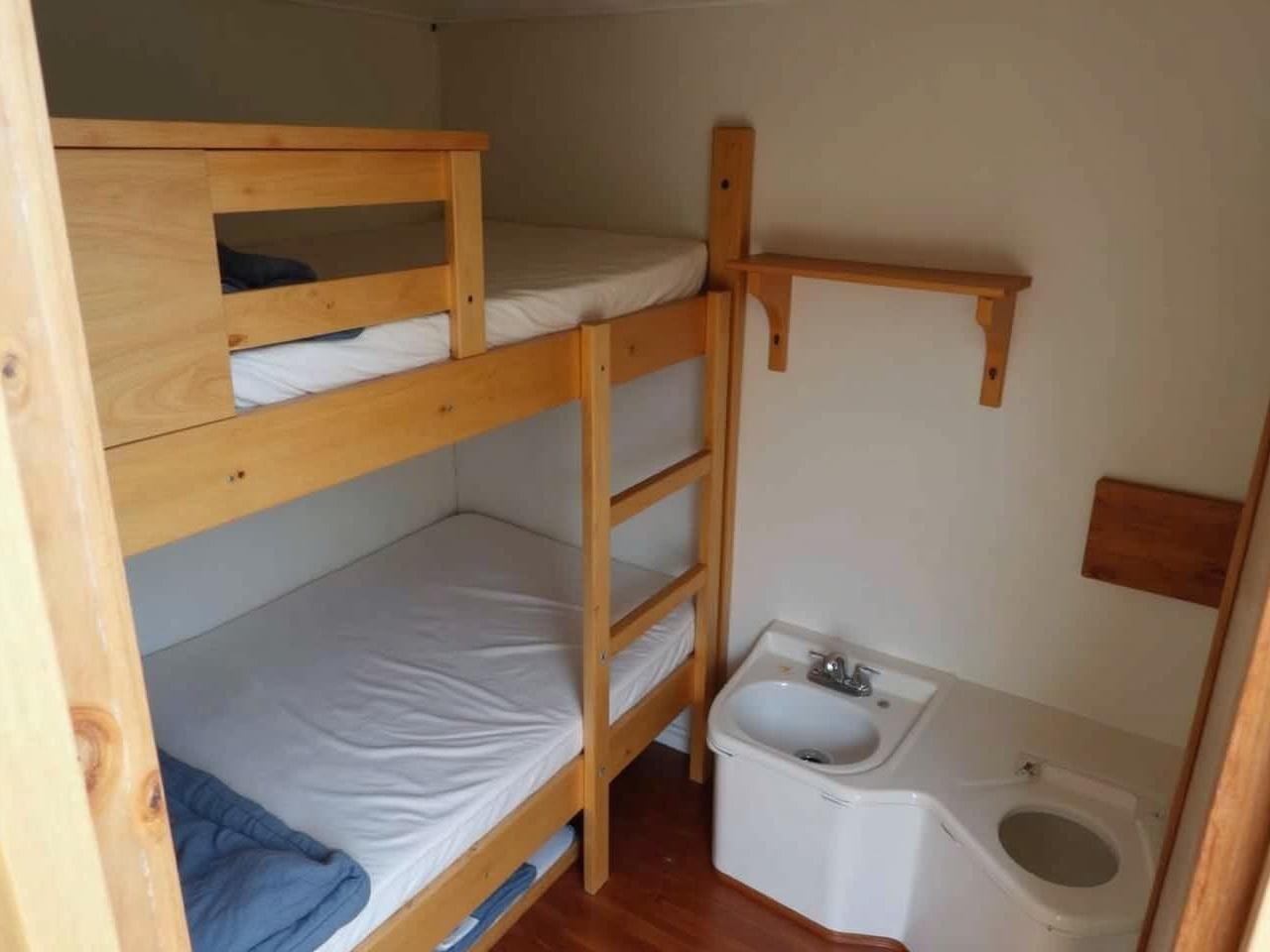 The image shows the interior of a small sleeping cabin or room. There are two stacked beds with a wooden ladder leading to the upper bunk. The bedding appears to be basic, with a mattress on each level. Next to the beds, there is a small area that looks like a storage shelf. In the corner, there is a compact washbasin or toilet area. The overall design seems functional and utilitarian, reflecting a practical usage of space.