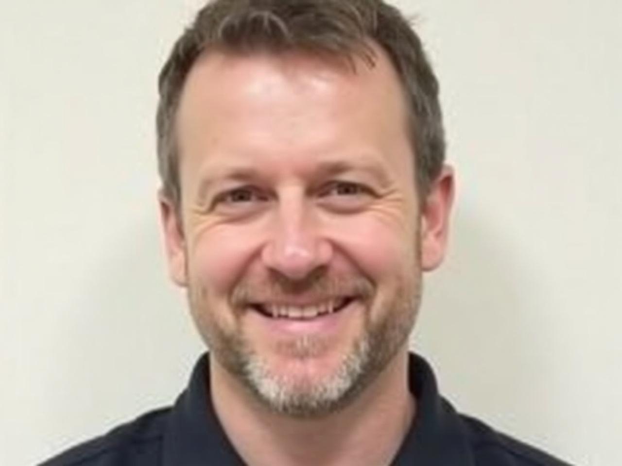 The image shows a man with a light beard and short, dark hair. He has a friendly smile and is wearing a dark shirt. The background is plain and light-colored, which helps the focus remain on his face. His expression appears warm and approachable, suggesting he may be someone who is easy to talk to. The lighting in the image is bright, emphasizing his features.