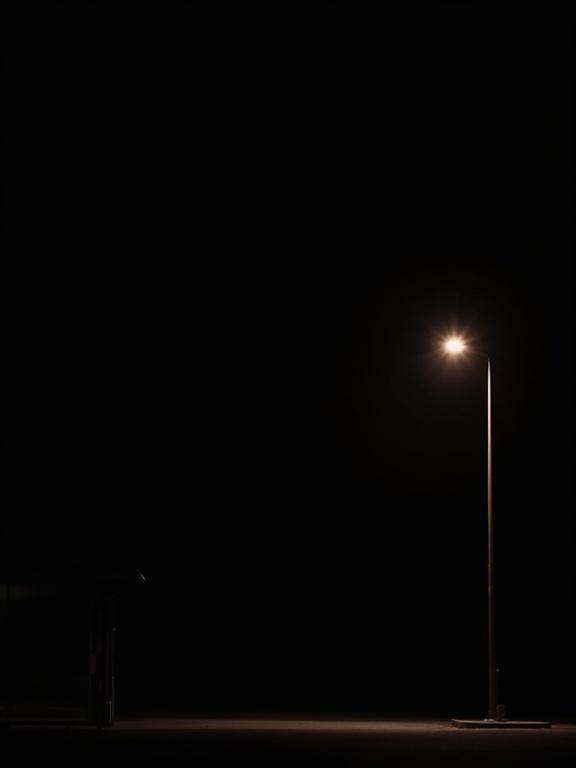 Silhouette of a tall street light at night with illumination near a bus stop placed at the bottom left. Large empty space fills the canvas with a black background.