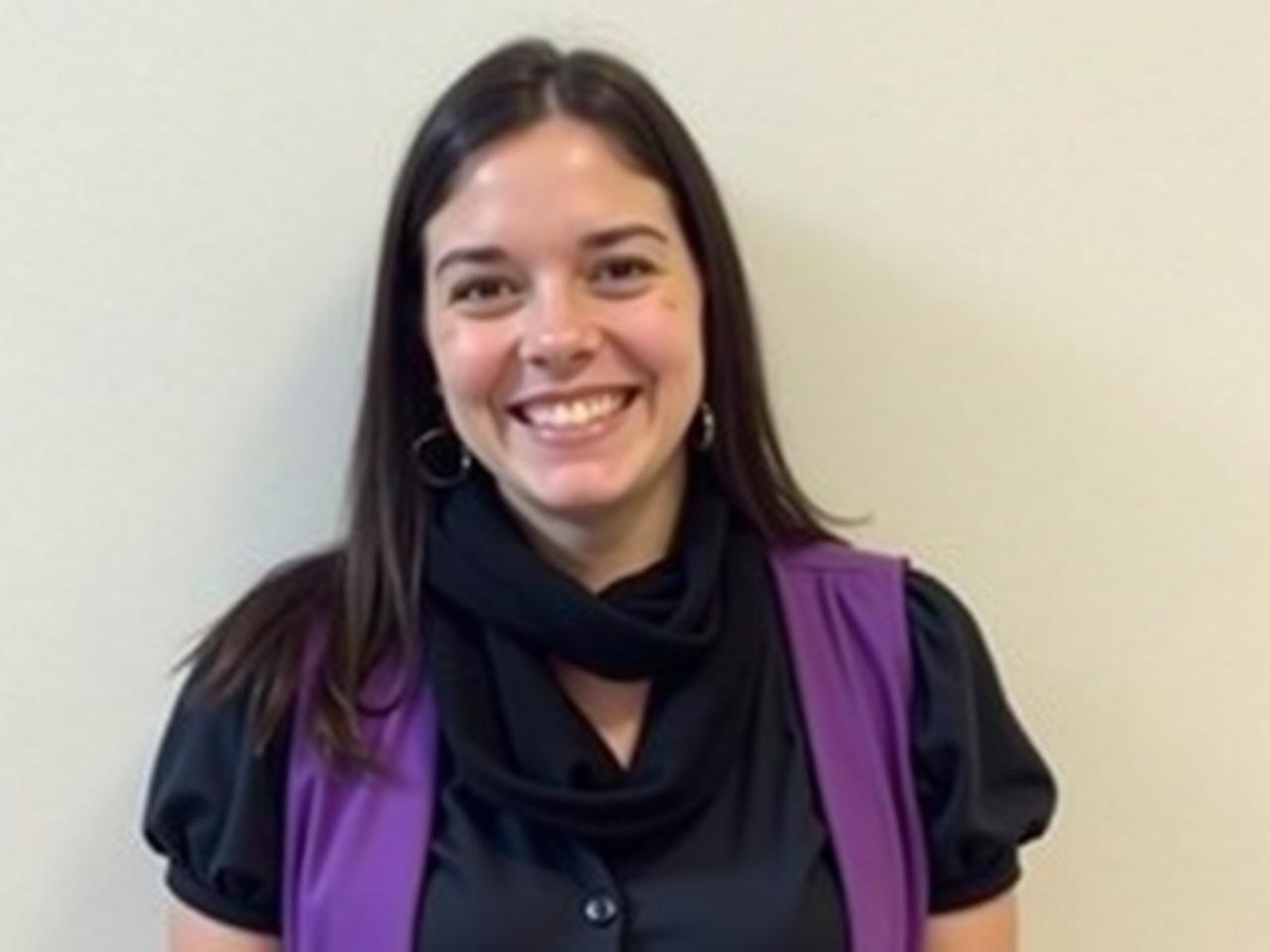 The image shows a woman smiling while standing against a light-colored wall. She is wearing a dark shirt with short puffed sleeves and a purple vest over it. The woman has long, dark hair and is accessorized with a black scarf around her neck. She appears to be in a casual setting, exuding a cheerful and friendly demeanor. The background is simple and does not distract from her presence in the photo.