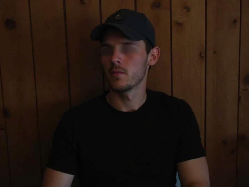 The image features a person sitting in front of a wooden wall. The person is wearing a dark cap and a plain black t-shirt. The lighting is dim, giving the photo a soft ambiance. There is a slight blur over the person's face to protect their identity. The background shows a simple wooden texture, enhancing a cozy atmosphere.