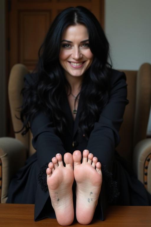 Mature goth woman has long black hair. Showing tattooed soles of bare feet. Sitting on a chair with feet on the coffee table. Smiling. Wearing black clothing.