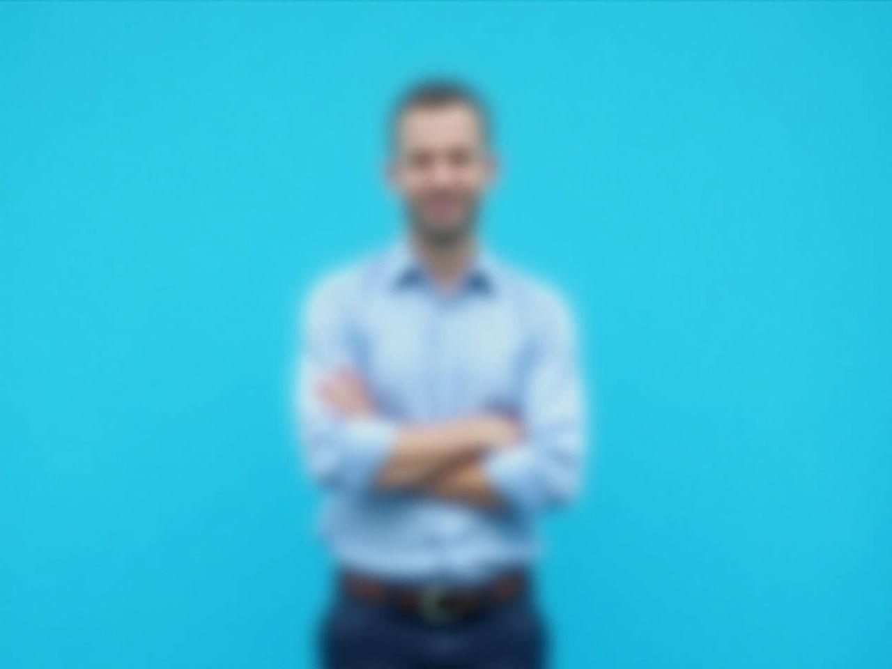 The image features a man standing against a bright blue background. He is wearing a light blue shirt with the sleeves rolled up and dark pants. His arms are crossed in front of his body, conveying a relaxed yet confident stance. The man is not fully visible due to a blurred face, which focuses attention on his attire and posture. The overall mood of the image is professional and approachable.