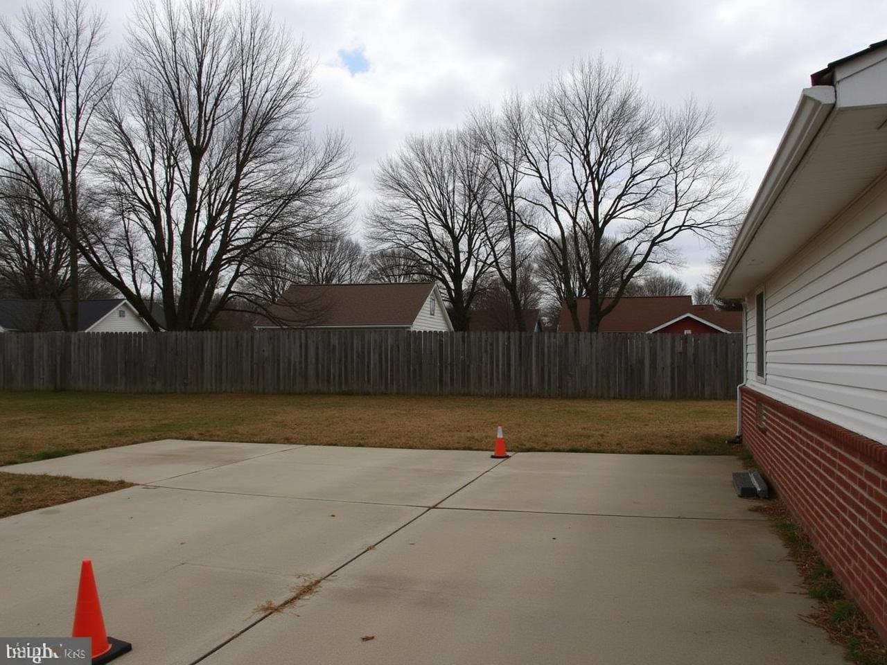 This image shows a residential area with a partially visible house on the right side. The house has a combination of white and brick exterior. In front of the house is an empty parking area with a subtle patch of what appears to be wet ground. To the left, there are two traffic cones placed on the ground. Behind the house, a wooden fence can be seen along with several trees that are apparently bare, indicating a possible autumn or winter season. The sky is covered with clouds, suggesting an overcast atmosphere.
