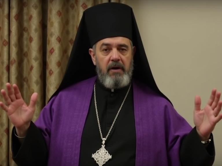 A religious figure delivering a speech. Middle-aged man with dark eyes and a graying beard. He wears a black pointed hood and a rich purple cape. Hands raised in a gesturing posture. Background has patterned drapes and light beige walls. Indoor lighting creates a warm atmosphere.
