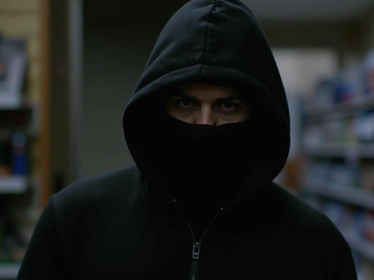 A person is seen wearing a black hooded jacket, with the hood pulled up over their head. The individual has their face mostly obscured, revealing only a small portion of their features. The setting appears to be indoors, likely a public space or store. The lighting is dim, making it difficult to see details clearly. The overall mood of the image suggests a sense of anonymity and secrecy.