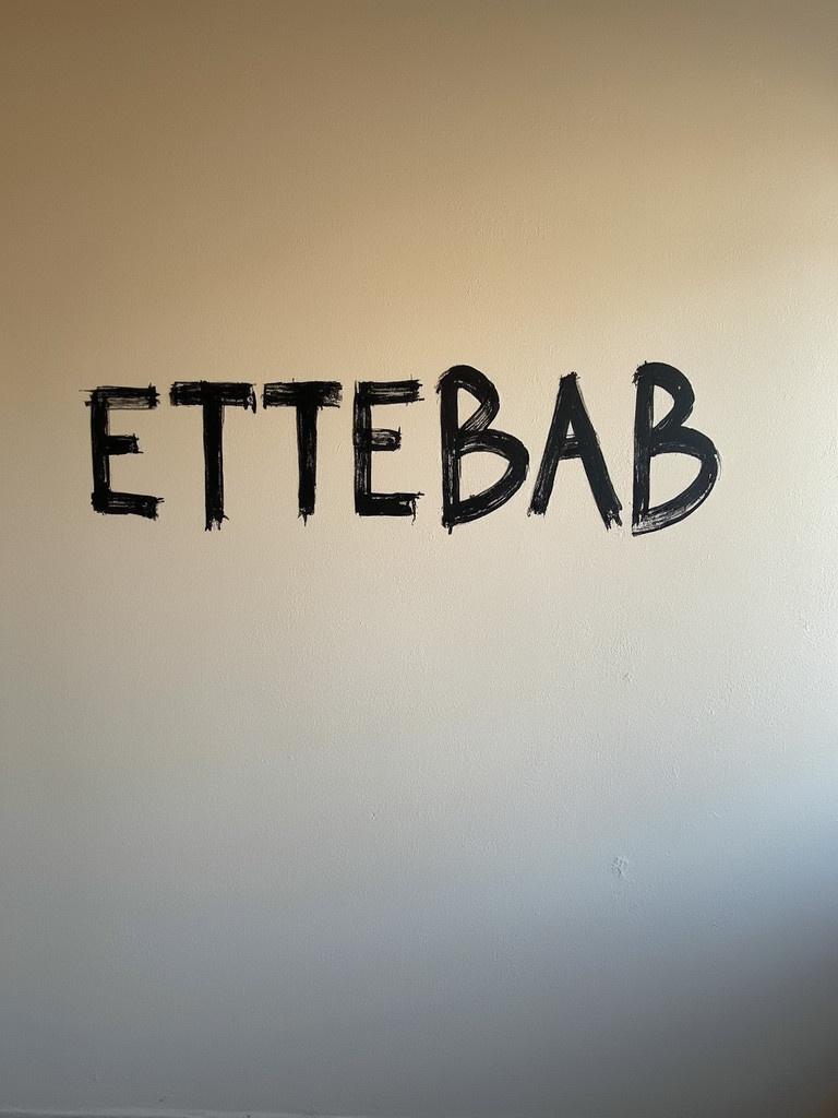 Wall painted with the name ETTEBAB in large letters. The letters appear bold and stylized. The background is a simple white. Lighting is soft and natural.