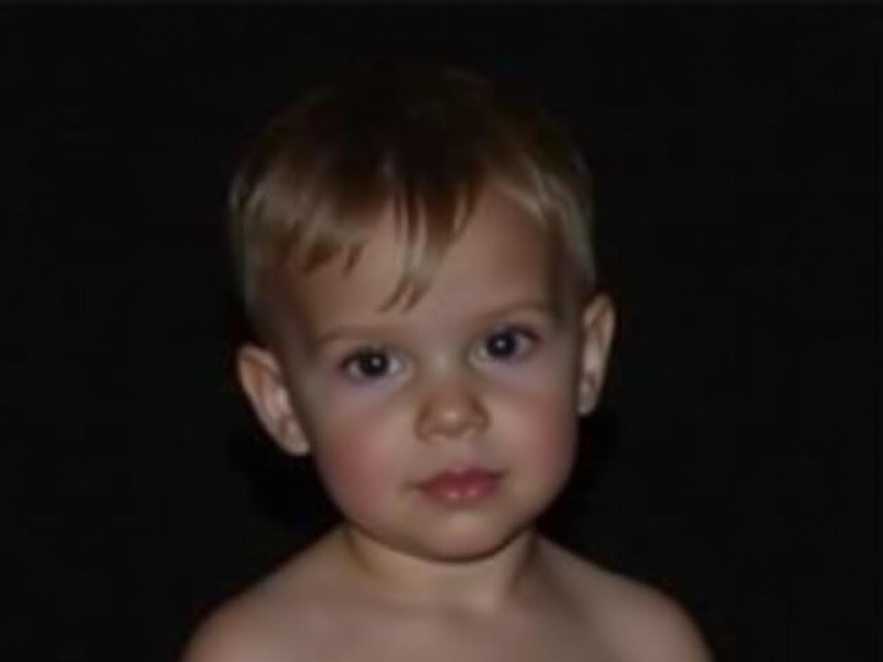 The image depicts a young child with a soft hairstyle, closely cropped around the sides with a fuller top. The child is facing forward with a neutral expression, their upper body visible. The background is completely dark, making the child stand out as the focal point. The child appears to be in a calm setting, possibly indoors, with soft lighting highlighting their features. There is a sense of innocence in the child's pose, contributing to an overall peaceful atmosphere.