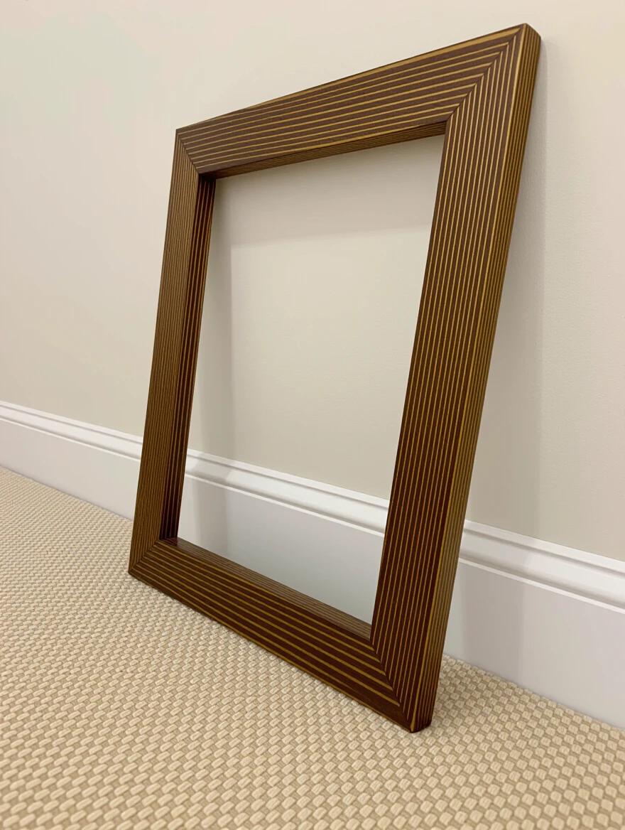 Wooden picture frame stands on the floor against a light colored wall. The frame has a width of 1 inch. A clean and simple design emphasizes the brown stripes of the frame.
