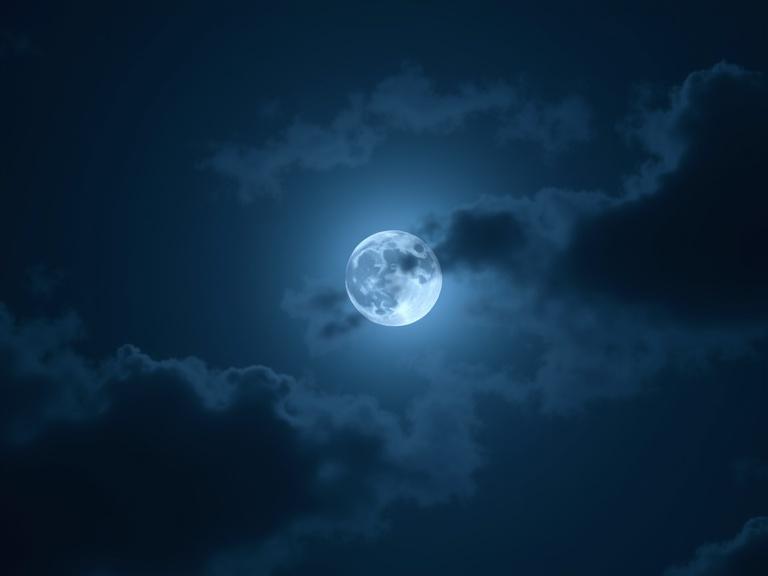 Clouds drifting across the moon in a dark night sky. Dim blue light creates a slow time-lapse feel. Displays haunting silence.