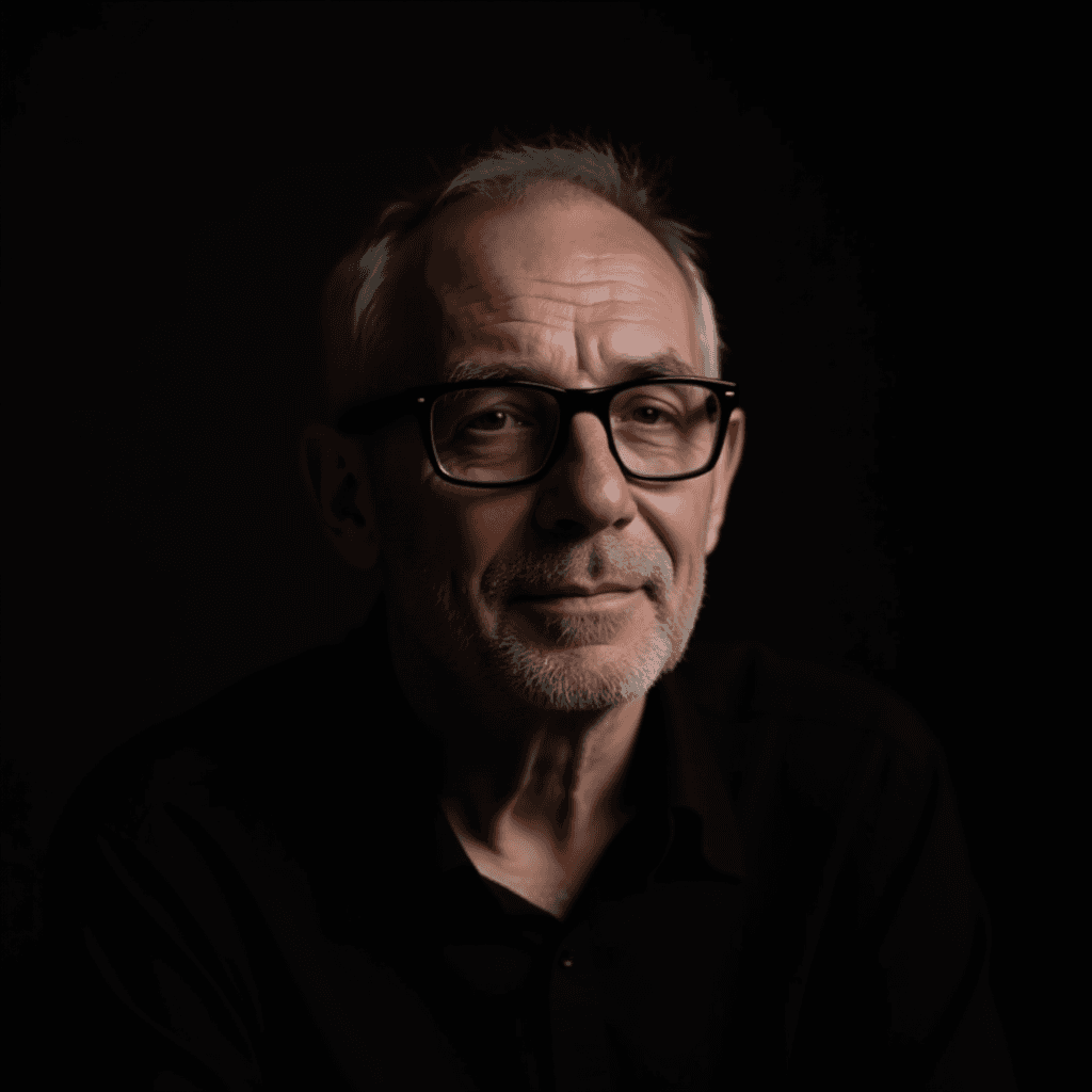 A man with glasses and a beard looks thoughtfully into the camera against a dark background.