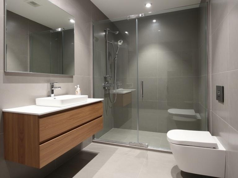 Modern bathroom features clean lines and neutral gray colors with warm wood accents. Large gray tiles cover walls and floor. There is a floating wood vanity with a white sink. A large frameless mirror is above the vanity. A walk-in shower has a frameless glass panel with chrome fixtures. A wall-mounted toilet is visible. Recessed lighting enhances the minimalist feel.