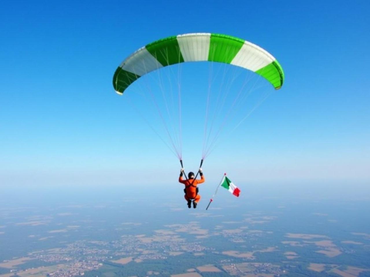 The image depicts a person skydiving with a parachute. The parachute has a vibrant design, featuring green and white sections. Below them, a landscape of fields and structures can be seen stretching out to the horizon. The sky is clear blue, indicating good weather for the jump. The person is wearing a bright orange jumpsuit and appears to be in a stable descent, skillfully controlling the parachute. Additionally, a small Italian flag is attached to the parachute, fluttering in the wind.