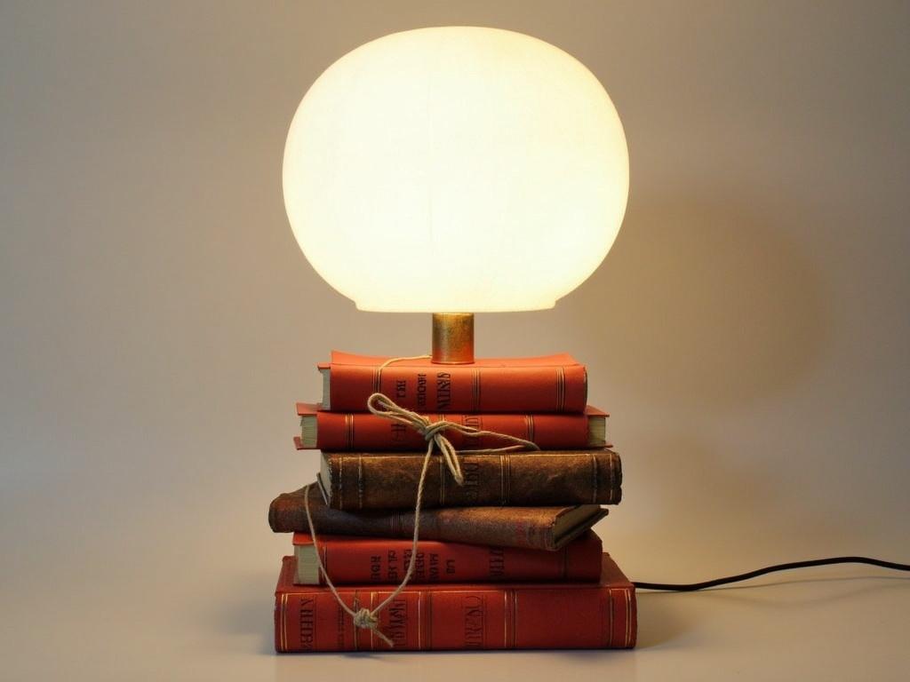 Imagine a stylish lamp designed to look like a stack of vintage books. The base is made up of several red and brown books, some tied together with string, while others are haphazardly placed. The books vary in size, creating a whimsical and eclectic look. Atop this unique base is a large, round, white lampshade that complements the colors below. The lamp emits a warm, inviting glow, perfect for reading or creating a cozy atmosphere. This piece would be a perfect addition to a home library or a reading nook.