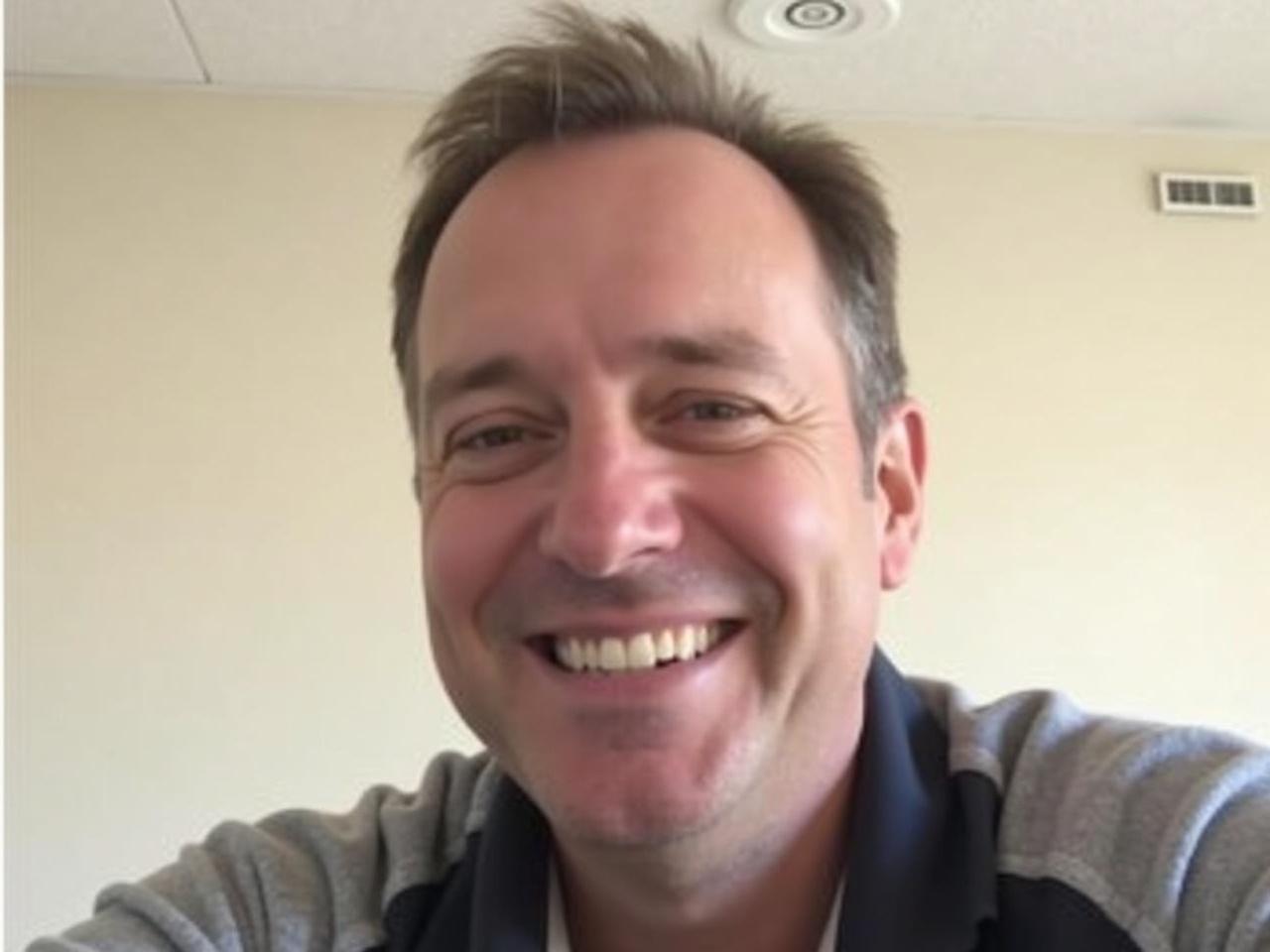 The image shows a man smiling for the camera. He has short, slightly wavy hair, and is wearing a dark-colored shirt with gray sleeves. The background is a light-colored wall, and there's a smoke detector visible on the ceiling. The lighting is bright, giving the image a cheerful vibe. The man appears relaxed and friendly.