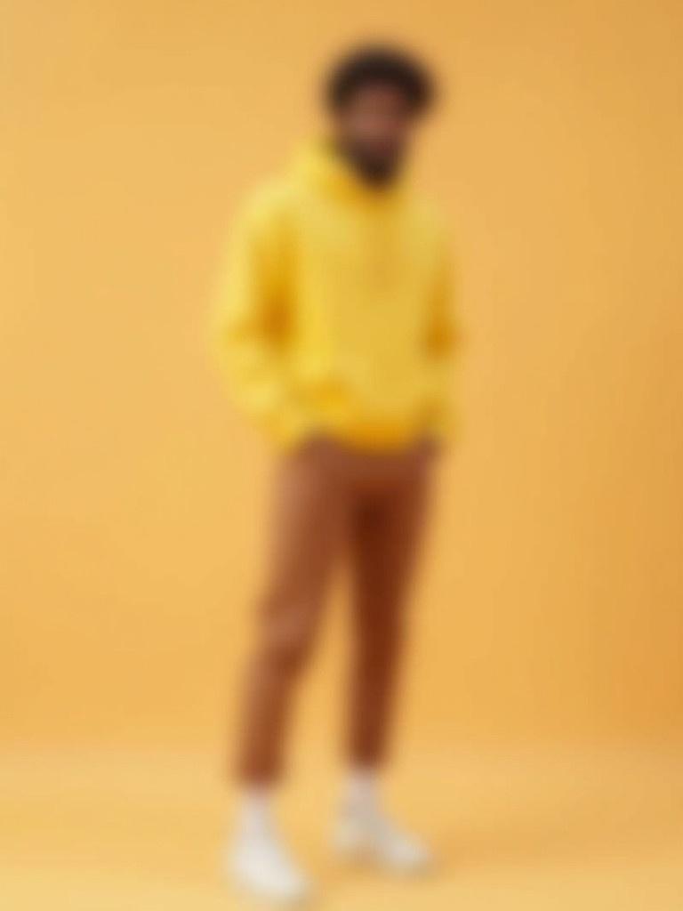 A man stands casually wearing a yellow hoodie and soft brown leggings. He has on white socks and shoes. The background is beige in color.