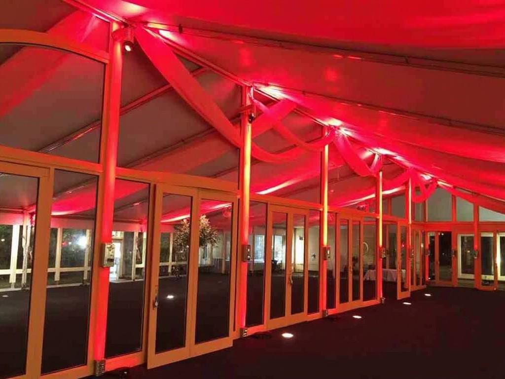 The image shows a spacious event tent with a bold red lighting scheme. The ceiling and walls are adorned with soft red fabric, creating a warm and inviting atmosphere. The floor is dark, emphasizing the illuminated features. Large glass doors provide access to additional spaces or outdoor views. This venue is ideal for various events, including weddings and corporate parties.