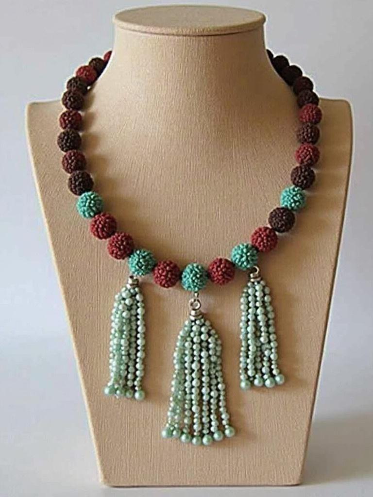 Colorful beaded necklace with tassels. Handmade from unique materials. Features round beads in red, brown, and teal. Decorative tassels made of smaller beads. Showcased on a bust display.