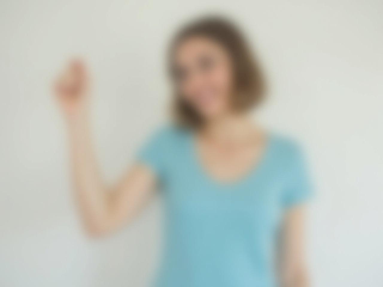 The image features a woman standing against a neutral background. She is wearing a light blue top, which is simple and elegant. The woman is raising her hand, but her hand is now a closed fist instead of an open hand. Her posture is confident, and she appears to be engaging with the viewer. The overall tone of the image conveys a sense of strength and assertiveness.