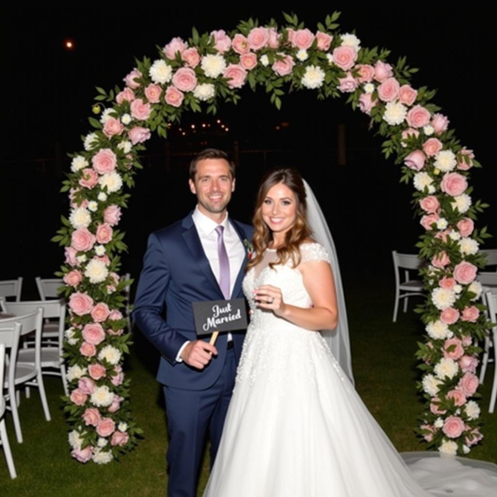 Newlywed couple stands in front of a floral archway. Groom wears dark blue suit. Bride is in white wedding dress with a veil. They hold a sign that reads Just Married. Floral archway has pink and white flowers. Chairs are set up for guests. Background is dark creating a romantic atmosphere.