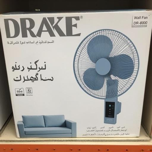 Box for a gray wall fan. Brand name DRAKE in bold letters. Wall fan model number DR-8000 displayed. Arabic lettering describes product features. Image shows light gray/blue fan with black control panel. Fan blades appear frosted. Light gray couch in lower left. Promotional text includes specifications like 18 inch. Image is well-lit with neutral background. Overall style is promotional and informative.