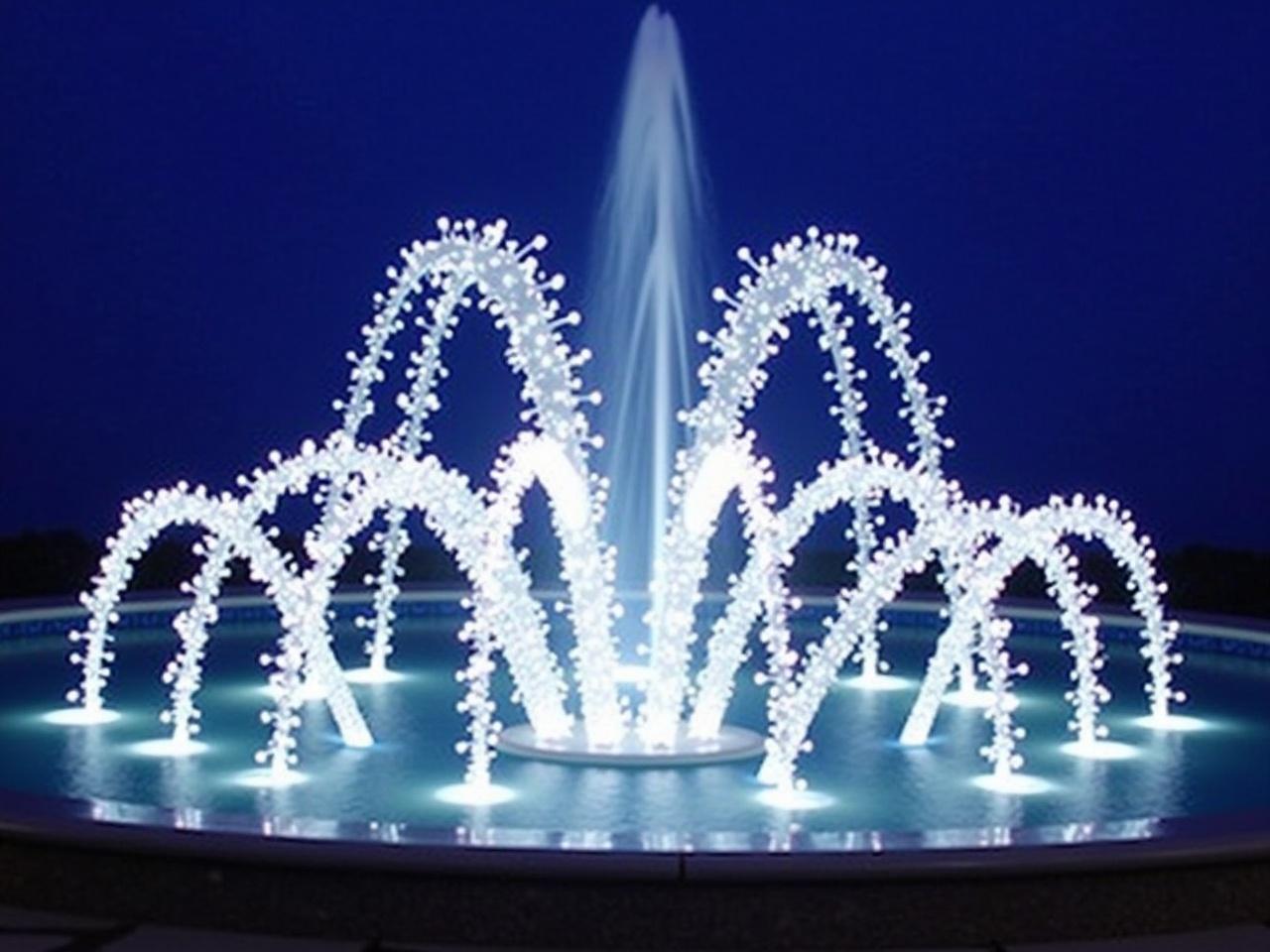 The image shows a beautiful, illuminated fountain. It has multiple arcs made of bright white lights. The background is a deep blue, enhancing the glow of the lights. The design is intricate, resembling a series of flowing streams that create an elegant display. The lights seem to twinkle, giving a festive and enchanting effect. This fountain is likely a decorative piece for an outdoor setting.