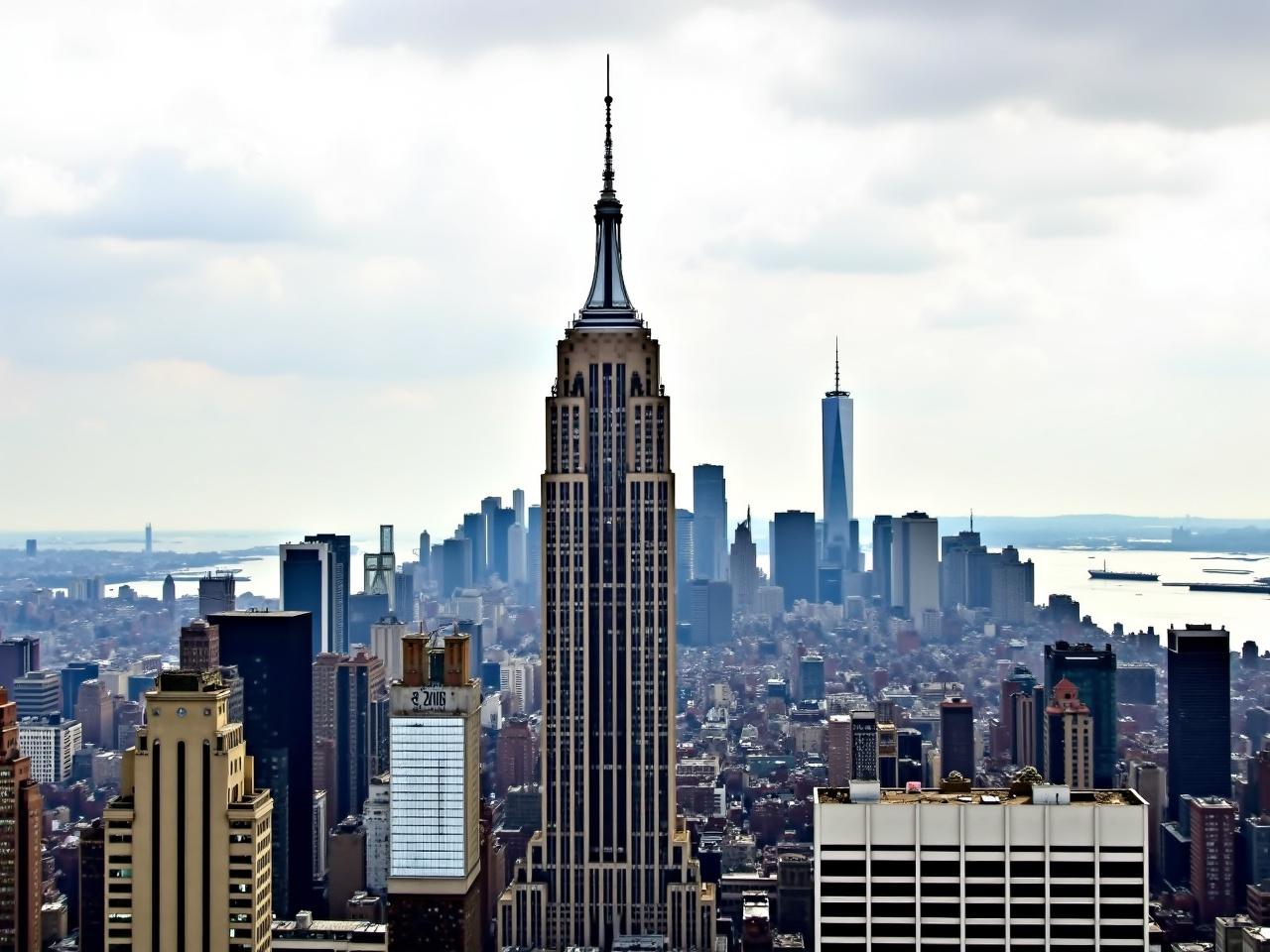 The image showcases a panoramic cityscape of New York City. In the foreground, the iconic Empire State Building stands tall with its distinct Art Deco architecture. Its spire is visible against a backdrop of clouds, suggesting a somewhat overcast day. Surrounding the Empire State Building are various other skyscrapers, including some with modern glass facades. In the distance, the One World Trade Center can be seen among other buildings. The cityscape hints at a vibrant urban environment full of architectural diversity.