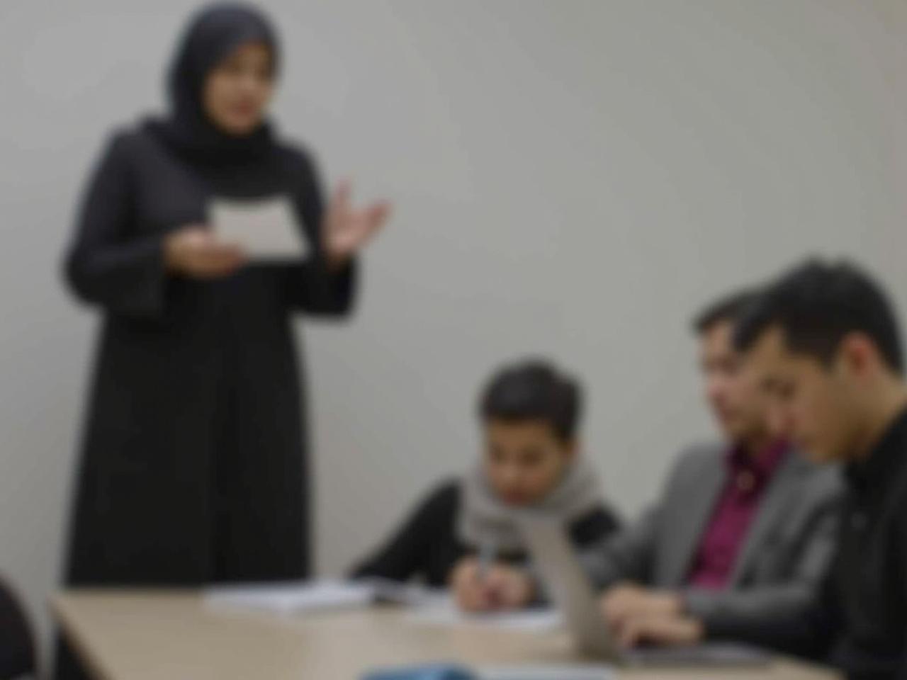 The image shows a person wearing a black hijab and a long black outfit while giving a presentation. They are standing and holding a piece of paper in one hand and gesturing with the other. The background is simple and soft, likely indoors, focusing on the speaker. There are two other individuals seated at a table in the foreground, engaged in the discussion. One person appears to be using a laptop, while the other is taking notes or interacting with materials on the table. The setting suggests a professional or educational environment.