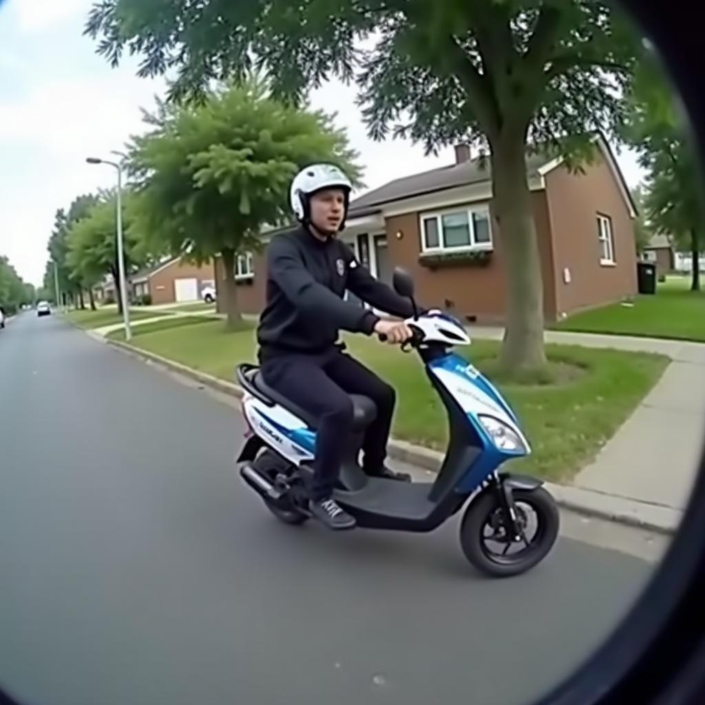 Bodycam footage shows a person riding a scooter on a street. The scooter is approaching a tree, indicating a possible crash. Street lined with trees and houses visible. Angle captures the scooter's side view.