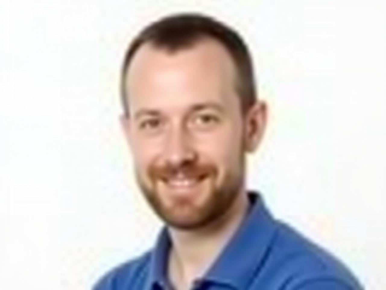 The image features a man with short, dark hair and a beard. He is wearing a blue polo shirt. The background is plain white, which helps to focus on his face. He has a neutral expression and is looking directly at the camera. The lighting is bright and even, highlighting his facial features. There are no distracting elements in the background, making it a clear portrait.