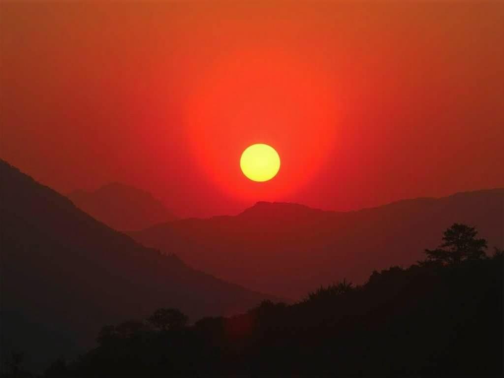 A stunning image capturing the sun setting over a range of mountains silhouetted against a bright red sky, conveying tranquility and natural beauty.