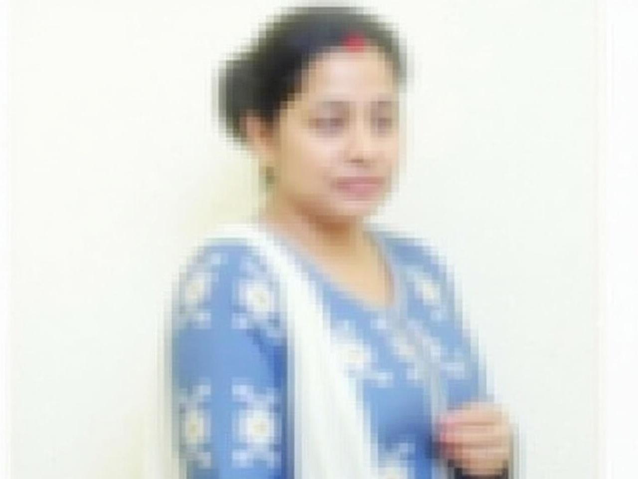 The image shows a woman with a blurred face, wearing a traditional outfit. She has a blue dress with white floral patterns and is draped in a white shawl. The woman has her head turned slightly to the side, presumably engaged in some conversation. Her hair is styled neatly, appearing shiny and well-kept. The background is plain and white, focusing attention on her attire and posture. Overall, the scene conveys a sense of grace and cultural attire.