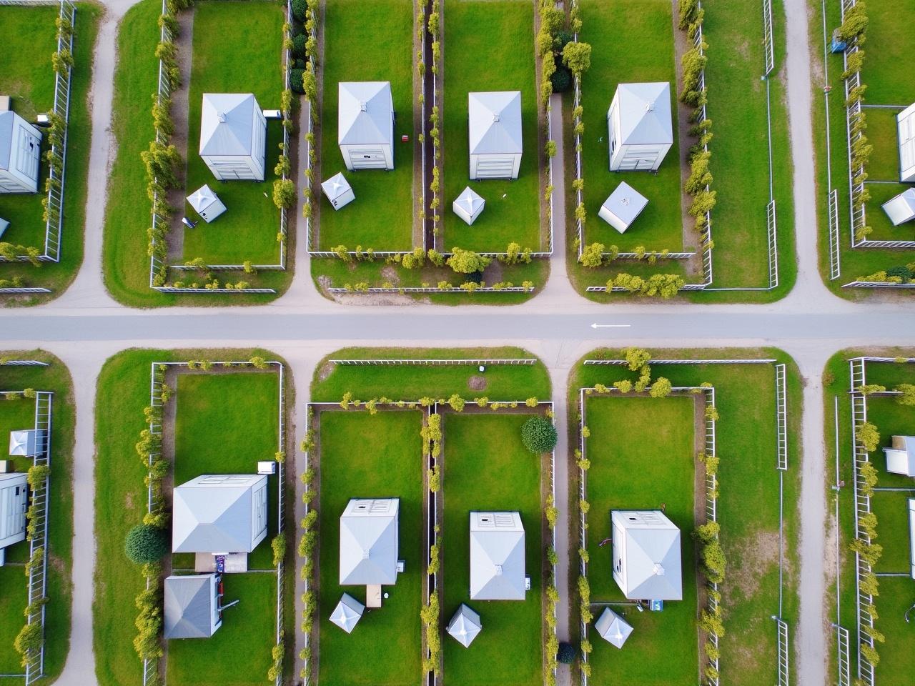 This is an aerial view of a neatly organized residential area. Multiple small, white houses are set within well-defined plots of land, each featuring a green lawn. The houses are arranged in rows alongside two parallel roads, allowing easy access to each property. Fences surround the plots, adding privacy and structure to the neighborhood. The area is spacious and clean, showcasing a well-planned layout with lots of green grass and pathways.