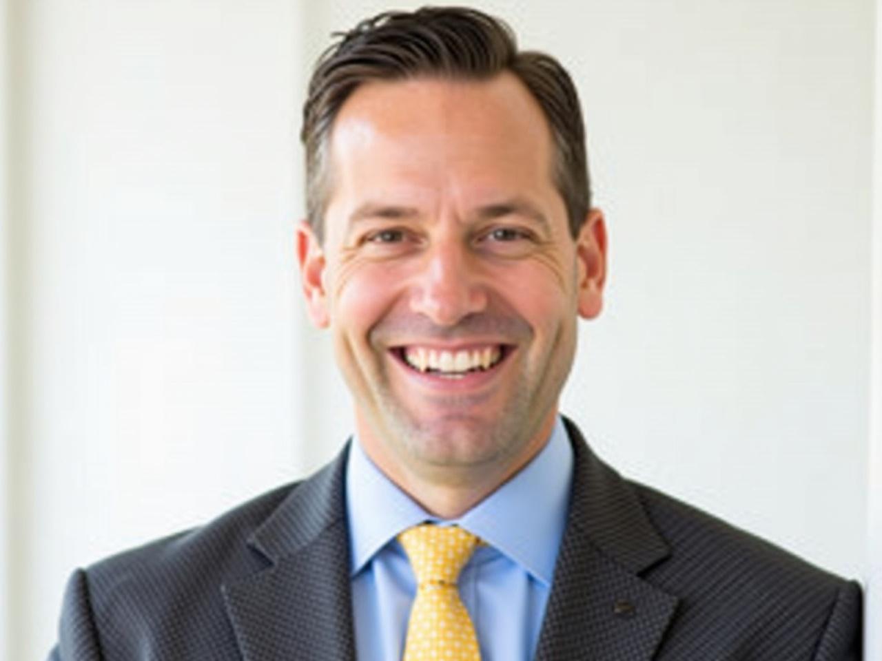 The image shows a man with a confident and friendly smile. He is wearing a dark, patterned suit and a light blue shirt, complemented by a yellow tie with small dots. The background is a simple, blurred white, which emphasizes his professional appearance. His hair is neatly styled, and he appears to be well-groomed. The overall vibe of the image is professional and approachable, making it suitable for a corporate or business setting.