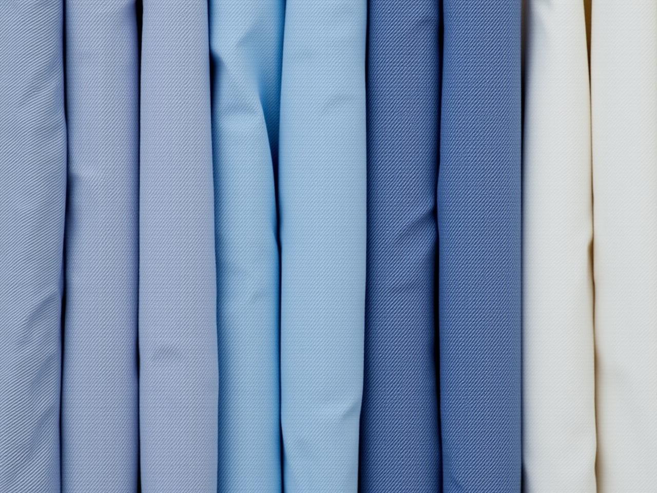 This image showcases a collection of folded shirts in various shades of blue and white. The fabrics are neatly organized, creating an appealing visual arrangement. The different tones of blue range from light sky blue to deep navy, complemented by crisp white. This presentation highlights the texture of the materials, inviting a sense of tactile interest. Overall, the soft lighting enhances the colors, giving the image a serene aesthetic.