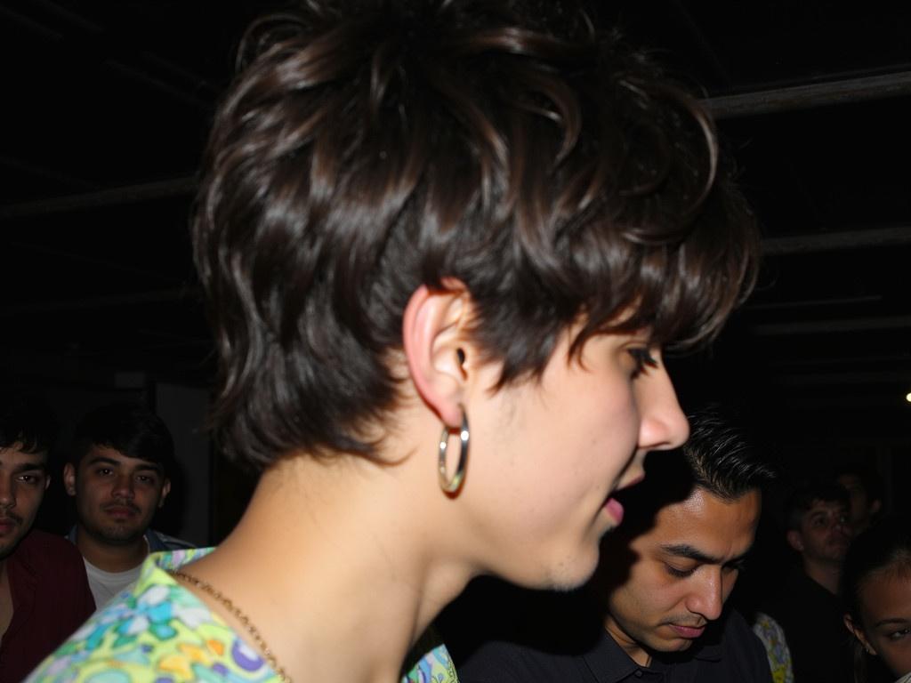 The image captures a side profile of a person's head, showcasing their hair and ear. The individual has dark, somewhat tousled hair that looks casual and unkempt. Their ear is adorned with a small hoop earring, adding a bit of personality. The shirt features a colorful, patterned design that can be glimpsed from the angle shown. The setting appears dimly lit, which creates an intimate atmosphere. This image gives a glimpse of a moment, possibly taken during a casual gathering or event.