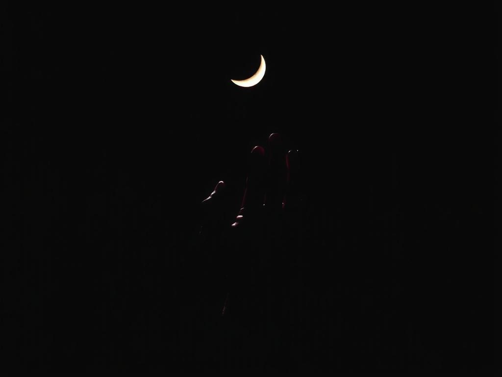 A silhouetted hand reaching towards a crescent moon in a dark sky.