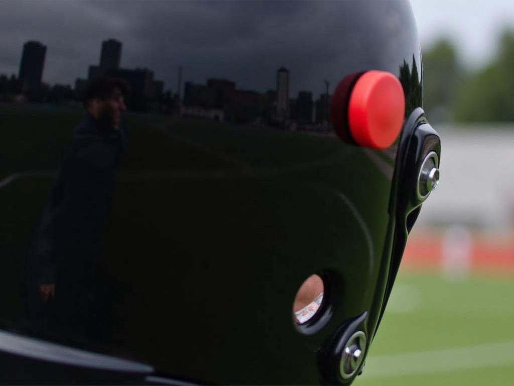 The image shows a close-up of a helmet. The helmet is primarily black with a shiny surface. There is a prominent red button positioned on the side of the helmet. The background is blurry, making it difficult to identify the surrounding environment. The overall focus seems to be on the helmet, hinting at themes related to sports or protective gear.