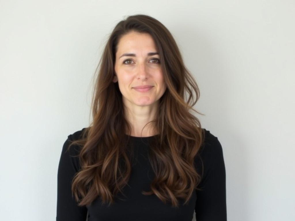 The image features a person standing against a plain background. They have long, wavy hair that falls freely around their shoulders. The individual is wearing a simple black top, which adds to the neutral tone of the image. The background is light-colored, providing a subtle contrast to their hair. The person appears to be looking directly at the camera, presenting a calm and composed demeanor.