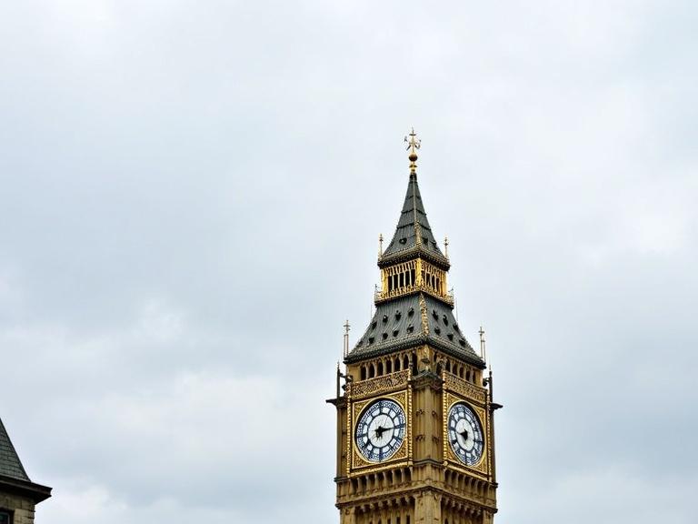 Photograph of Elizabeth Tower in London showcasing detailed architecture. Low angle captured cloudy sky. Gold accents, white clock faces with Roman numerals. Gothic style with stone texture. Centered in frame with slight shadow.