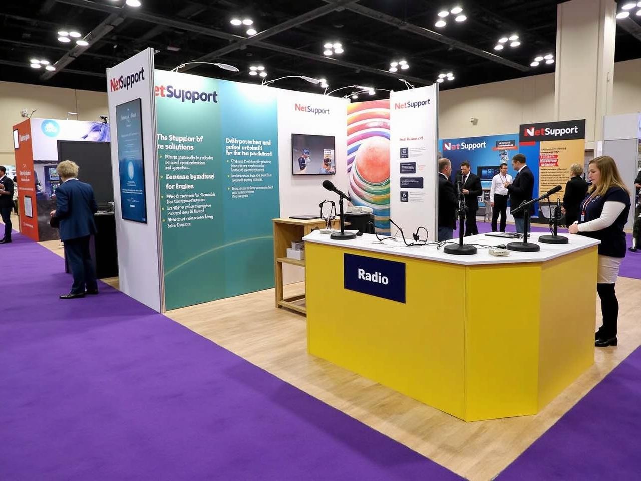 The image showcases a trade show booth for a company named NetSupport at an exhibition. The booth is vibrant and colorful, with various information panels highlighting their solutions for school networks and classroom management. There is a yellow station labeled 'Radio' with microphones and headsets for broadcasting, indicating an interactive element. The floor is covered in purple carpet, adding to the lively atmosphere. Surrounding booths display products and services related to education technology, creating a collaborative environment at the event.