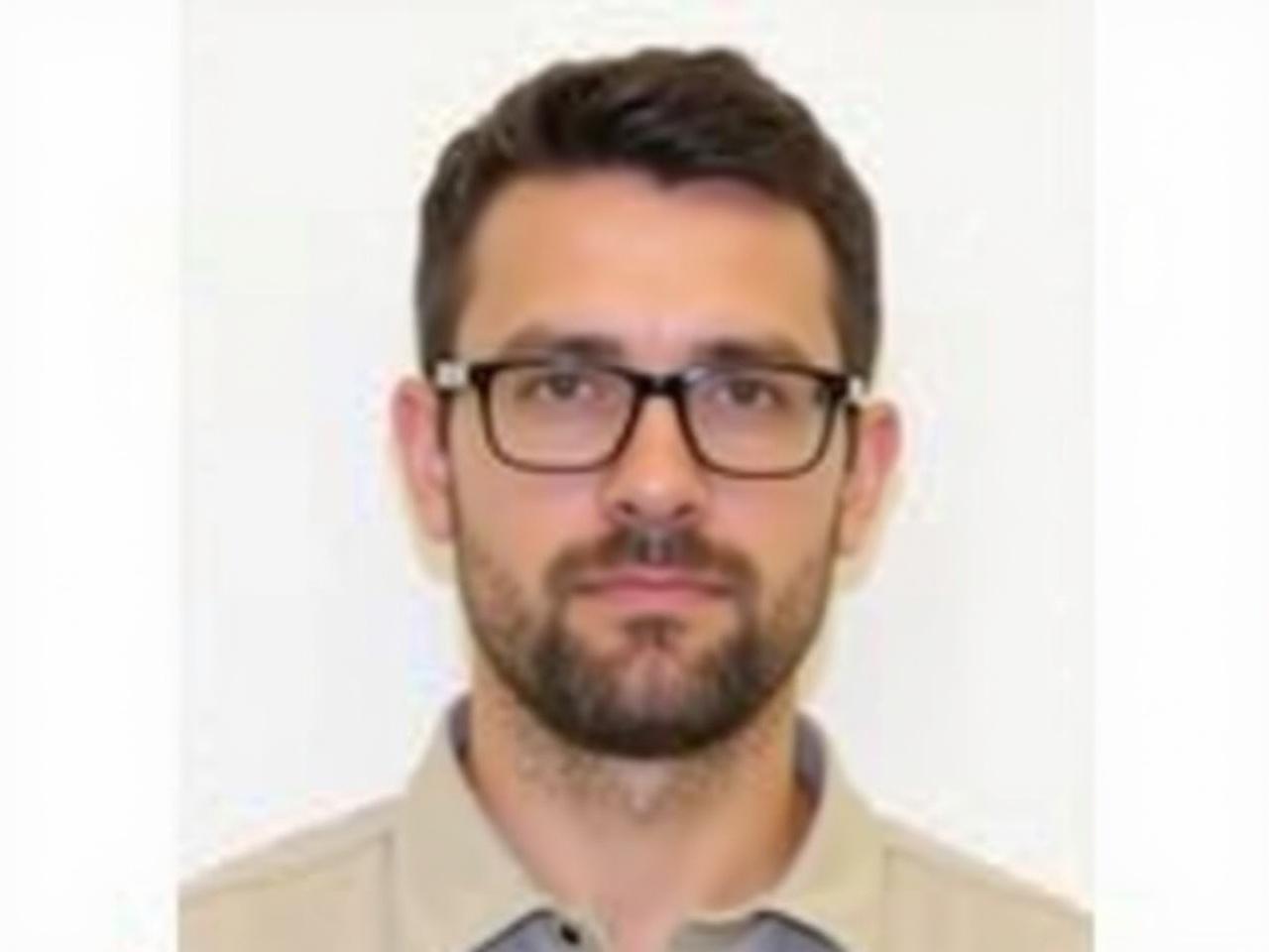 The image depicts a man with short, dark hair and glasses. He has a neatly groomed beard and is wearing a light-colored collared shirt. The background is plain white, ensuring that the focus remains on his face. He appears to have a serious expression, looking directly at the camera. The lighting is soft, highlighting his facial features clearly. Overall, the image conveys a professional and composed demeanor.