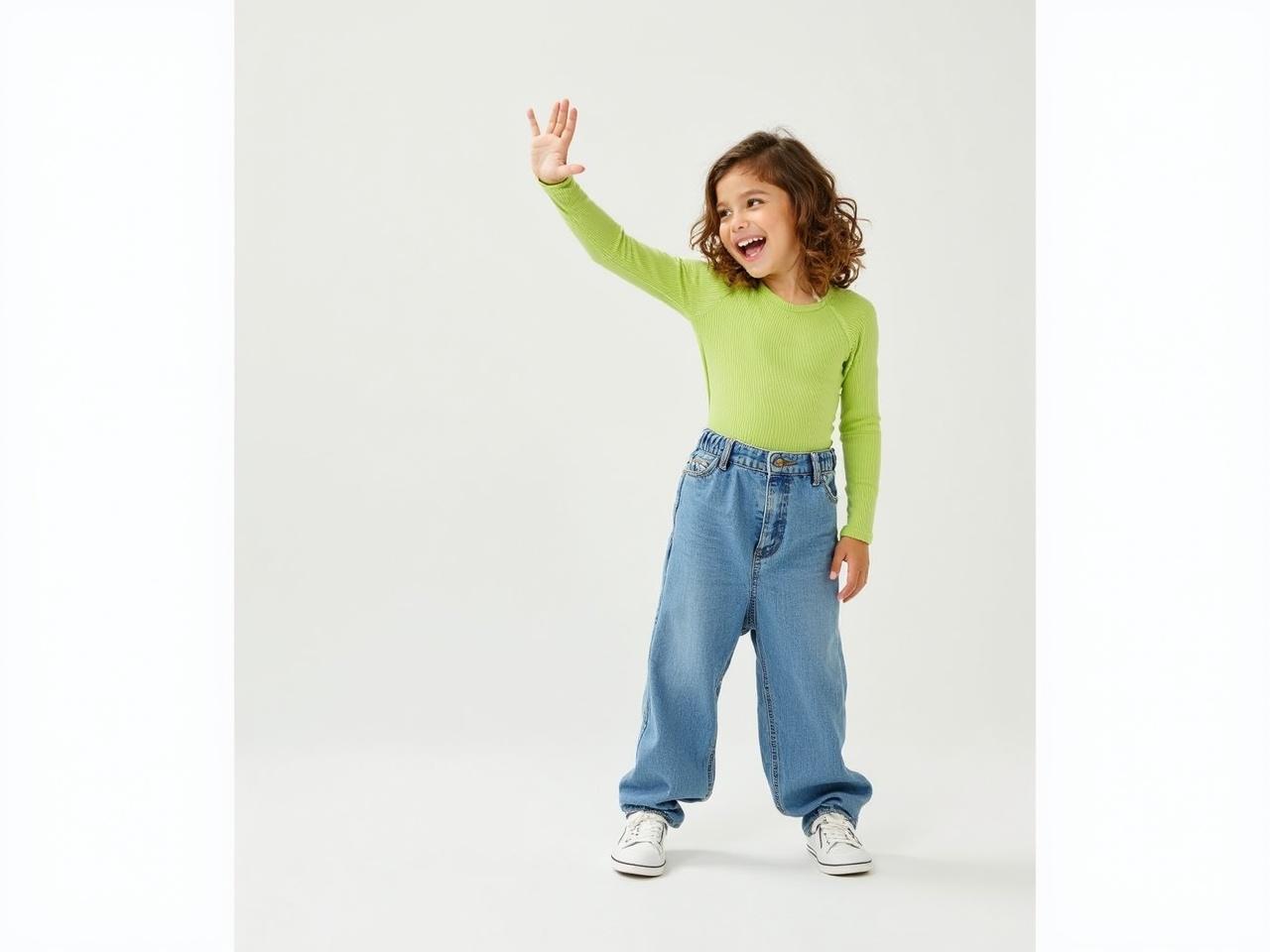 The image features a child posing against a white backdrop. They are wearing a vibrant green ribbed long-sleeve shirt and baggy blue denim pants. Their outfit is completed with white sneakers. The child has long, wavy hair. They are making a playful gesture with their left hand while their right arm is extended to the side.