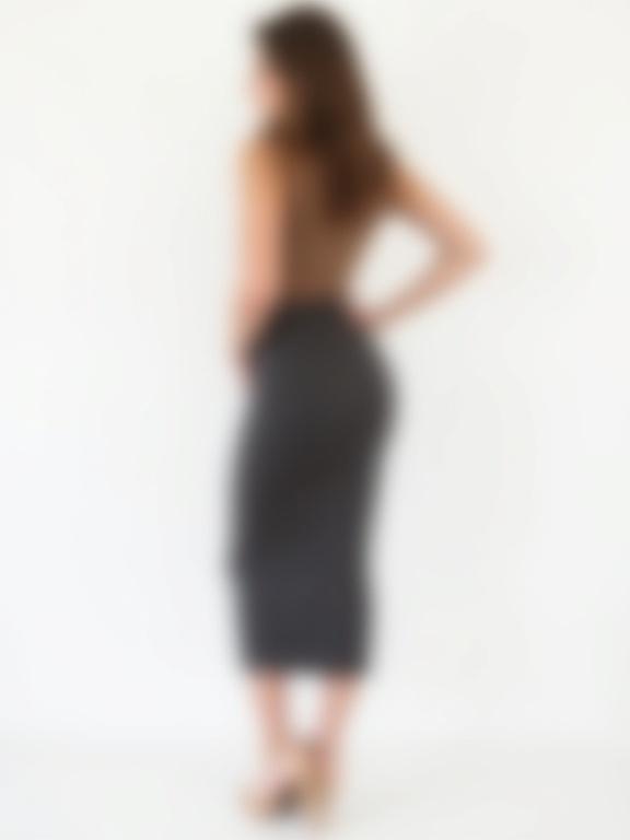 Image of a woman from behind in three-quarter view. Long wavy brown hair. Wearing a sleeveless brown or tan top and a long dark charcoal pencil skirt. Skirt is high-waisted and form-fitting. High-heeled nude shoes with ankle straps. One hand at her waist and looking over her shoulder. Plain white background with soft lighting creating subtle shadows.