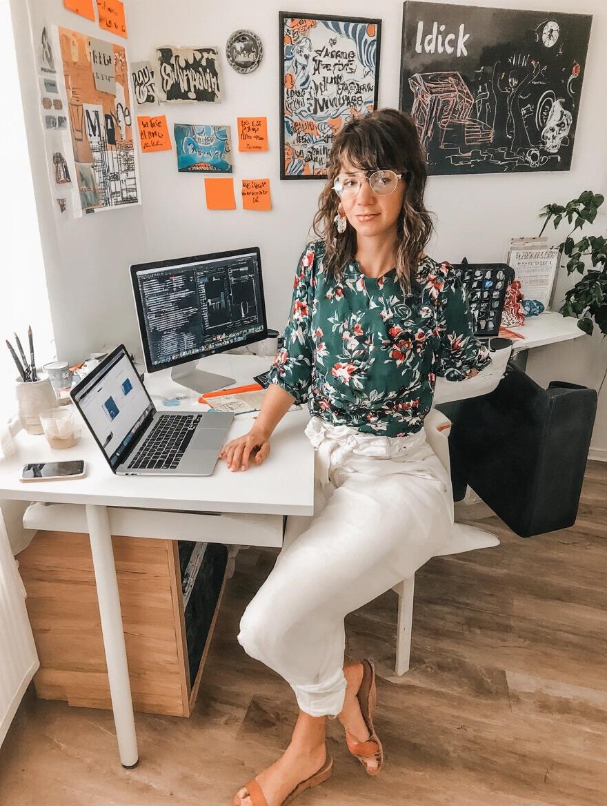 A woman sitting barefoot at her work desk. She has long hair and wears glasses. The desk has two computer screens and various decorations. A plant is visible nearby.