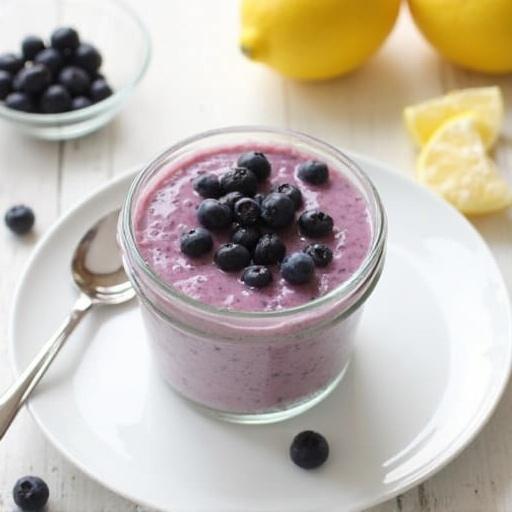 A vibrant blueberry lemon chia pudding placed in a clear jar. Topped with fresh blueberries. Surrounded by lemon slices and a small bowl of blueberries. A silver spoon beside the jar on a white plate.