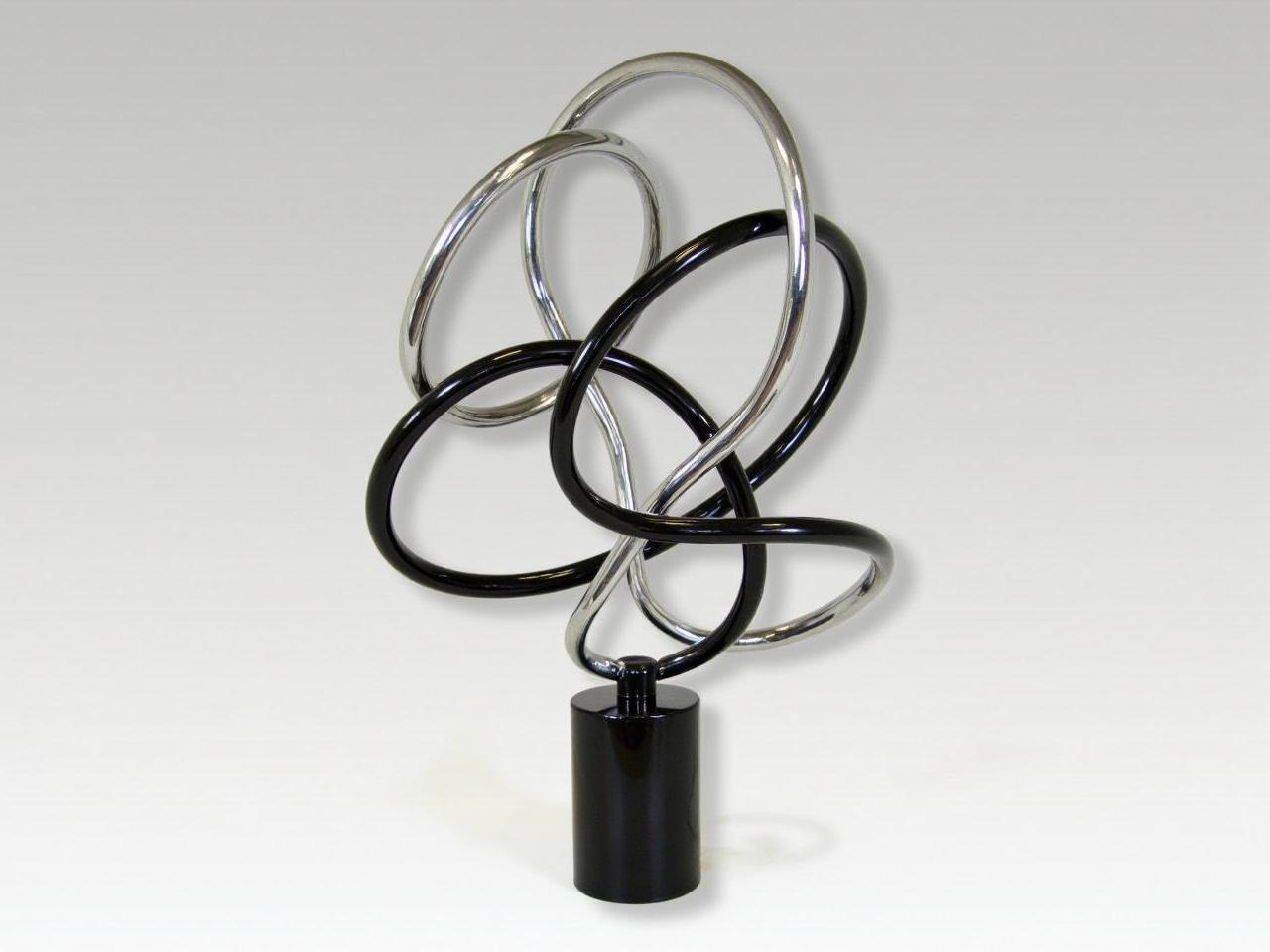 This image depicts a modern abstract sculpture made from metal. The sculpture features intertwined loops that vary in color, combining both silver and black finishes. It has a smooth, polished surface that reflects light, enhancing its aesthetic appeal. The base of the sculpture is sturdy and has a cylindrical shape that supports the complex structure above. The overall design appears dynamic, as if it is in motion, inviting viewers to admire it from various angles.