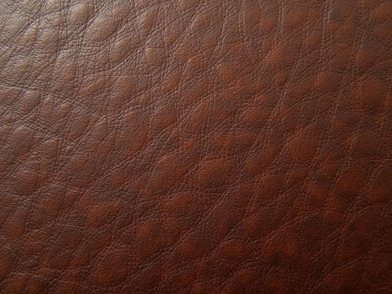 A close-up of micro leather grains in rich brown color. The texture appears detailed and smooth with visible patterns. The surface has a soft sheen and looks luxurious. Suitable for high-definition uses.
