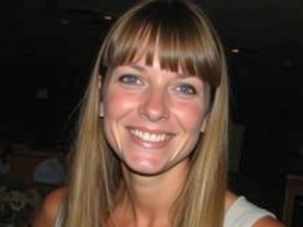 The image shows a woman smiling warmly at the camera. She has long, straight hair that falls past her shoulders with bangs framing her face. Her skin tone appears fair and she has bright blue eyes. The background is dark, possibly indicating an indoor setting, and there is a soft focus to the surroundings. The woman is wearing a light-colored top and appears to be in a joyful mood, conveying a sense of friendliness.