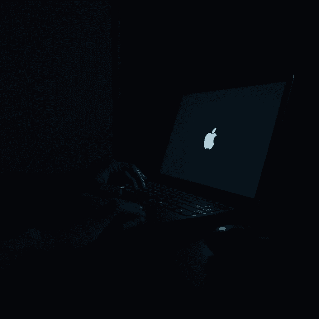 A person is using a laptop with a glowing Apple logo in a dark room.